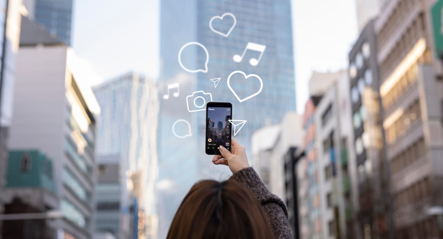 A person holds a smartphone with social media icons like hearts, music notes, and chat bubbles floating around it, set against a city backdrop.